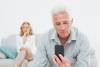 Senior man reading text message with displeased woman sitting on sofa in a house